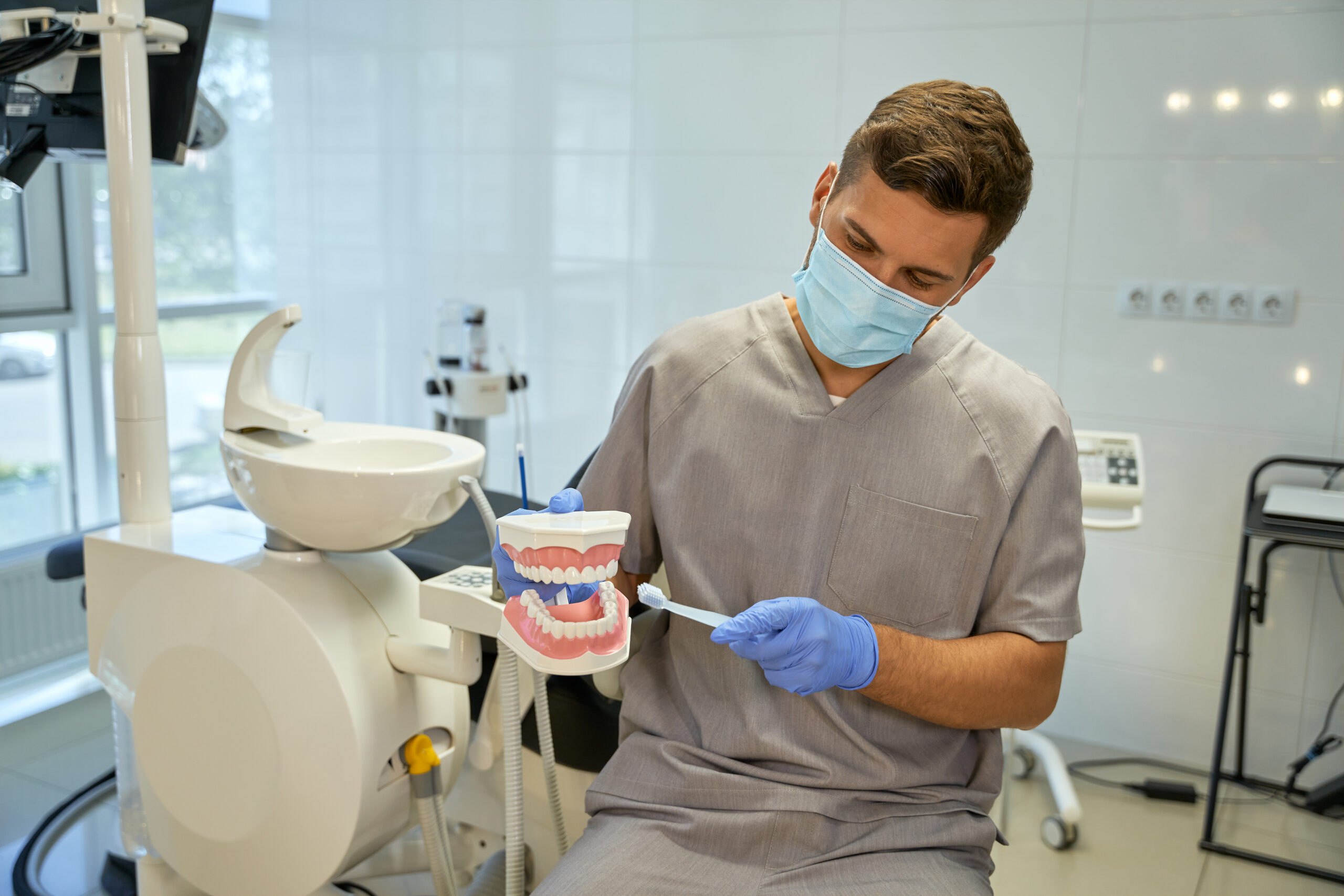 Caring doctor demonstrating proper way to brush teeth Professional dentist in a mask holding replica of jaws and a toothbrush while imitating brushing process